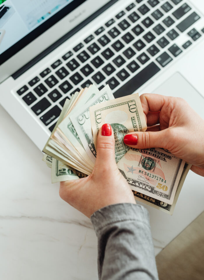 Person with red nail polish counting U.S. dollar bills in front of an open laptop on a white desk.