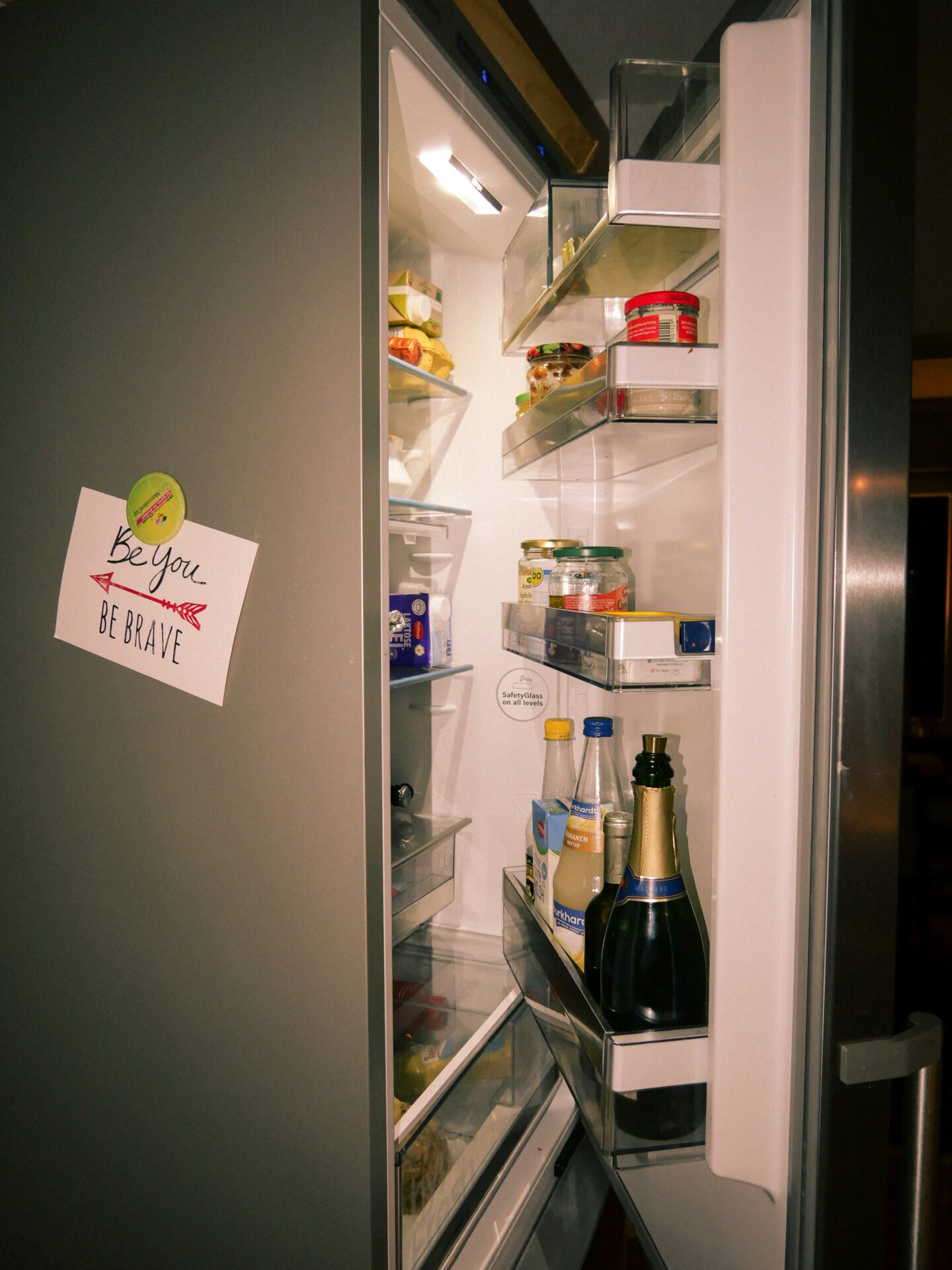 Open refrigerator in a kitchen filled with food, drinks, and condiments, with a motivational note on the door that says “Be you. Be brave.”