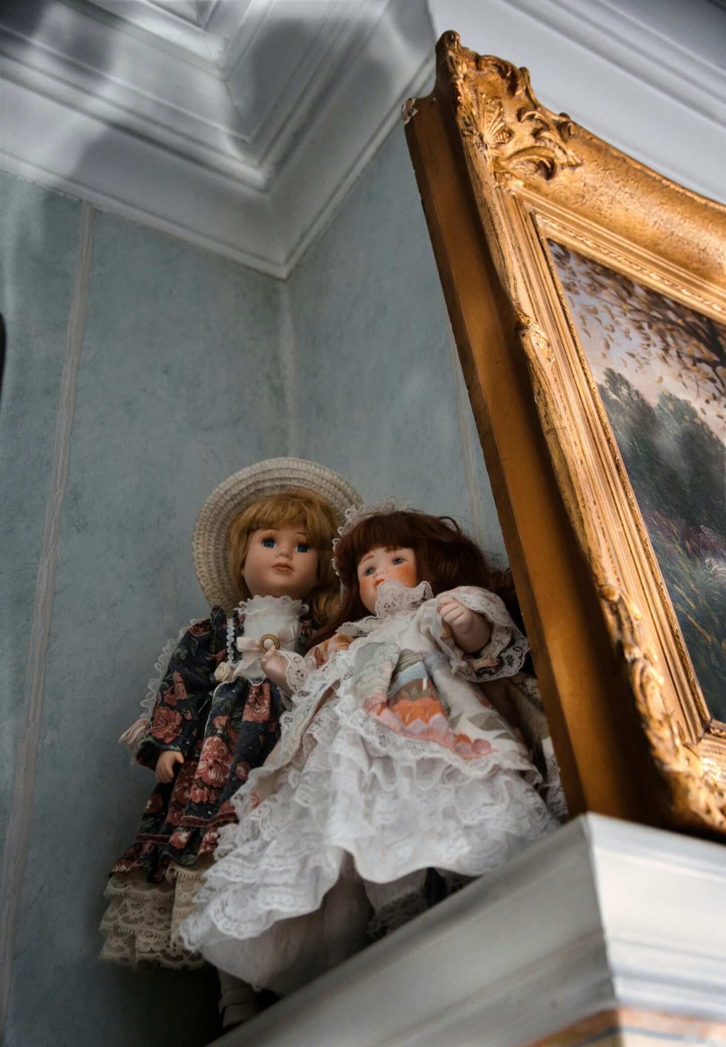 Two vintage heirloom quality dolls dressed in lace and floral dresses displayed on a shelf beside an ornate gold-framed painting.