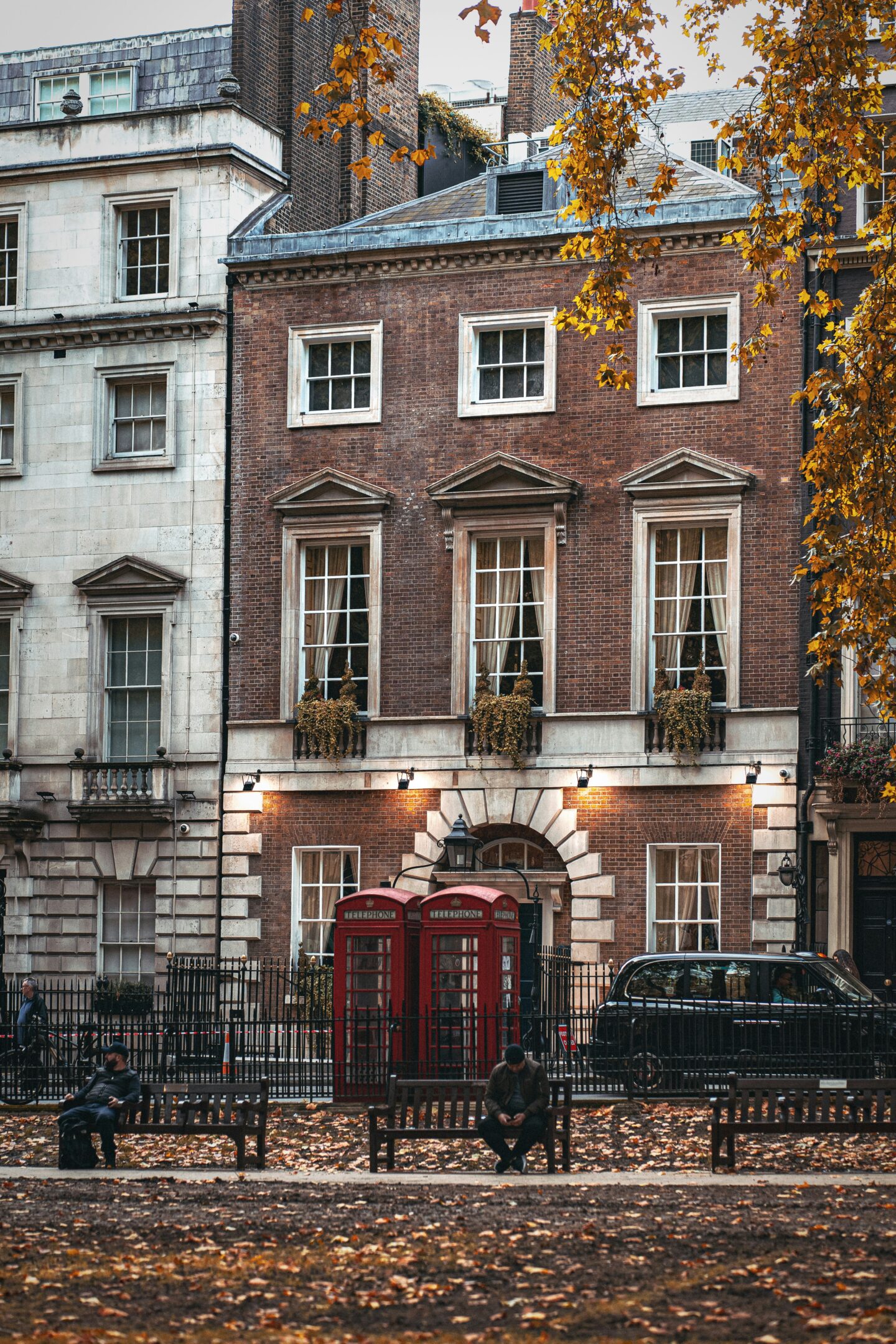 Charming London street in autumn with red telephone boxes, historic Georgian buildings, and fallen golden leaves in the park.