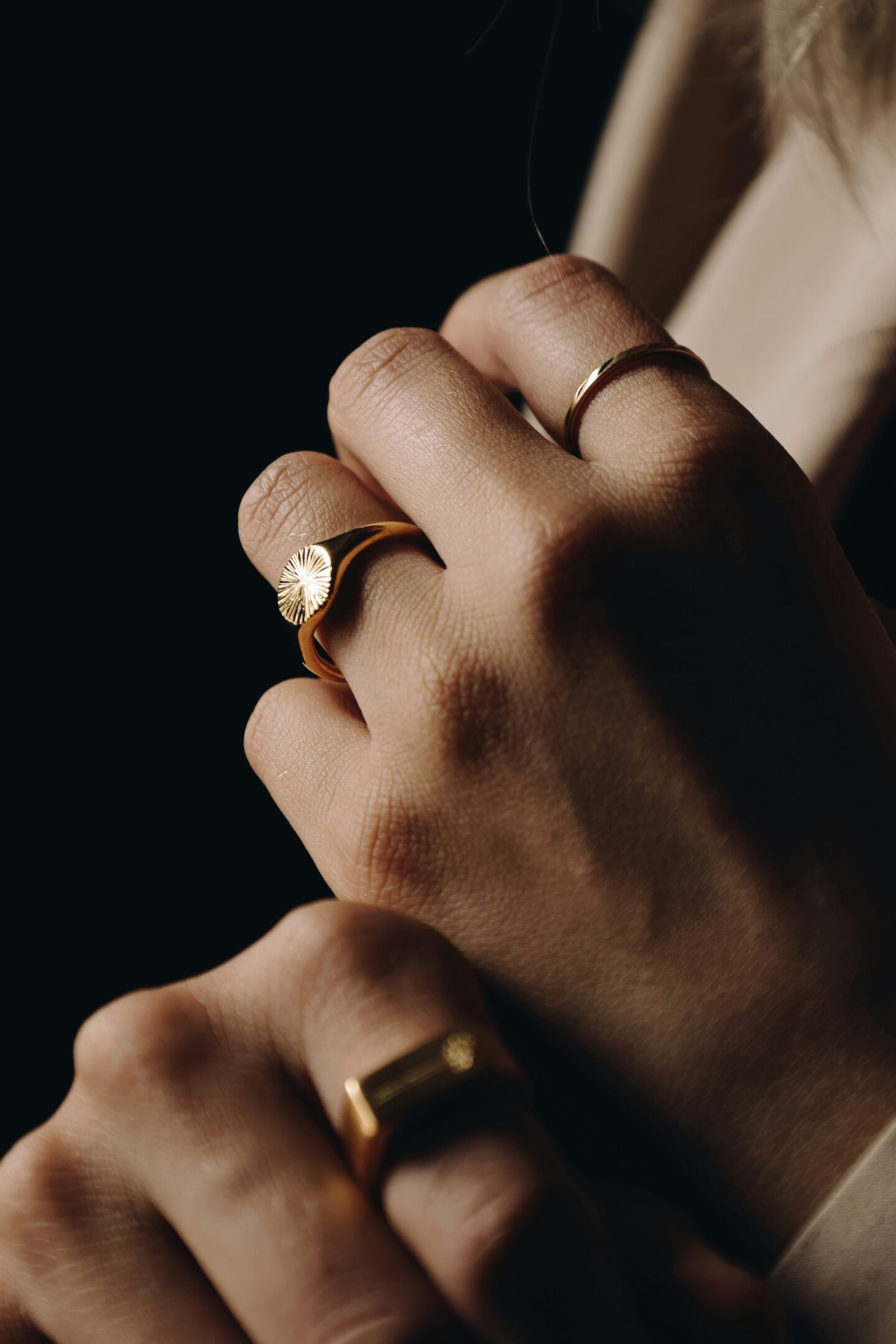 Close-up of hands adorned with gold rings, including a sunburst design signet ring, against a dark background.