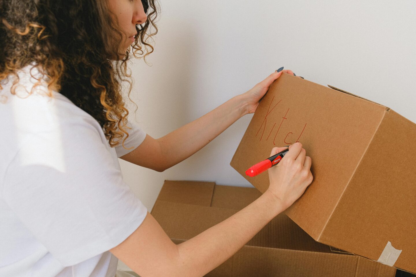 Woman labeling a cardboard box with the word “kitchen” using a red marker while packing for a move.