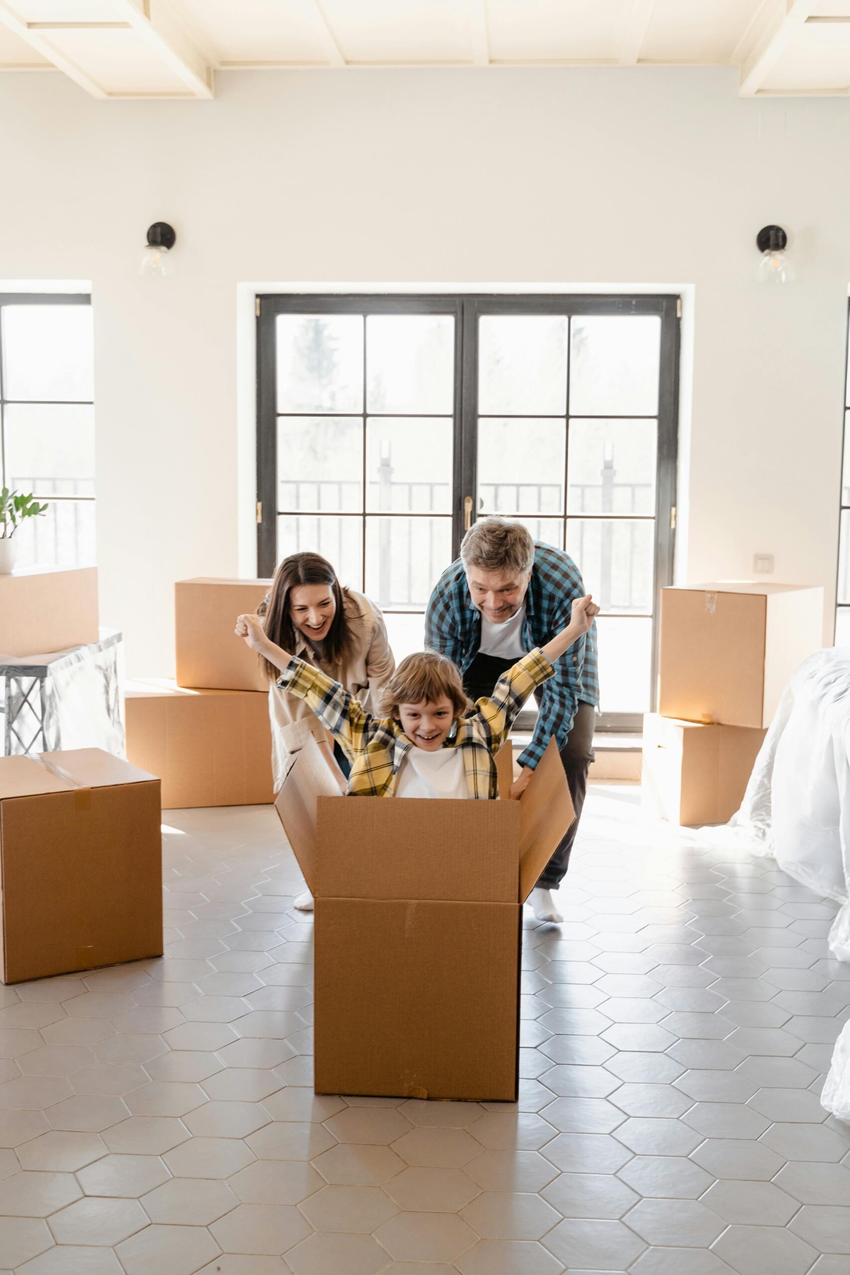A cheerful family playing with moving boxes in a bright room, with a child sitting inside a box and raising their arms excitedly.