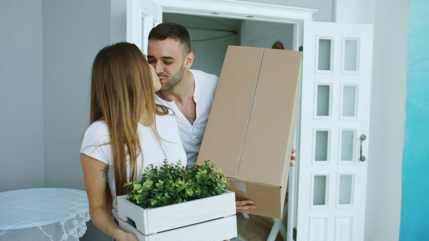 A couple kisses while carrying items into a house, the man holding a large cardboard box and the woman holding a white crate with green plants.