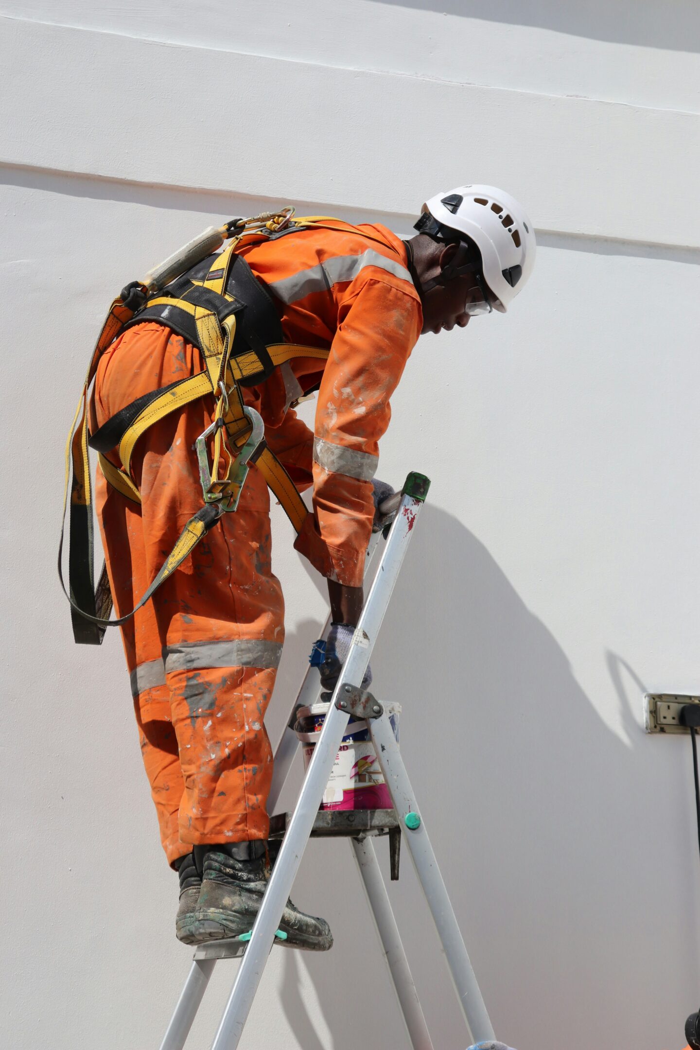Worker in an orange protective suit and safety harness painting a white exterior wall while standing on a ladder.