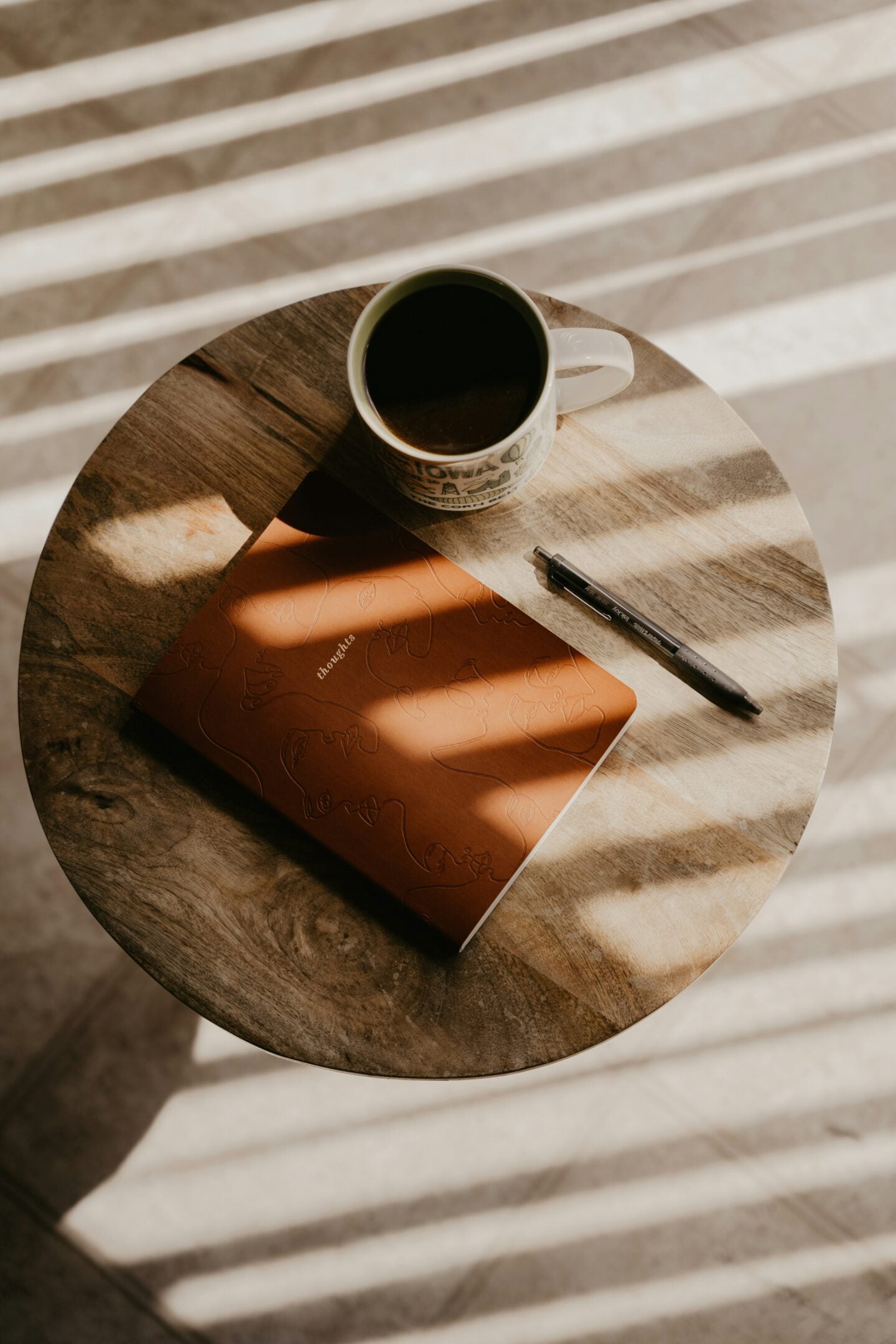 A cup of coffee, a pen, and a brown journal with an embossed design rest on a round wooden table with sunlight casting striped shadows across it.