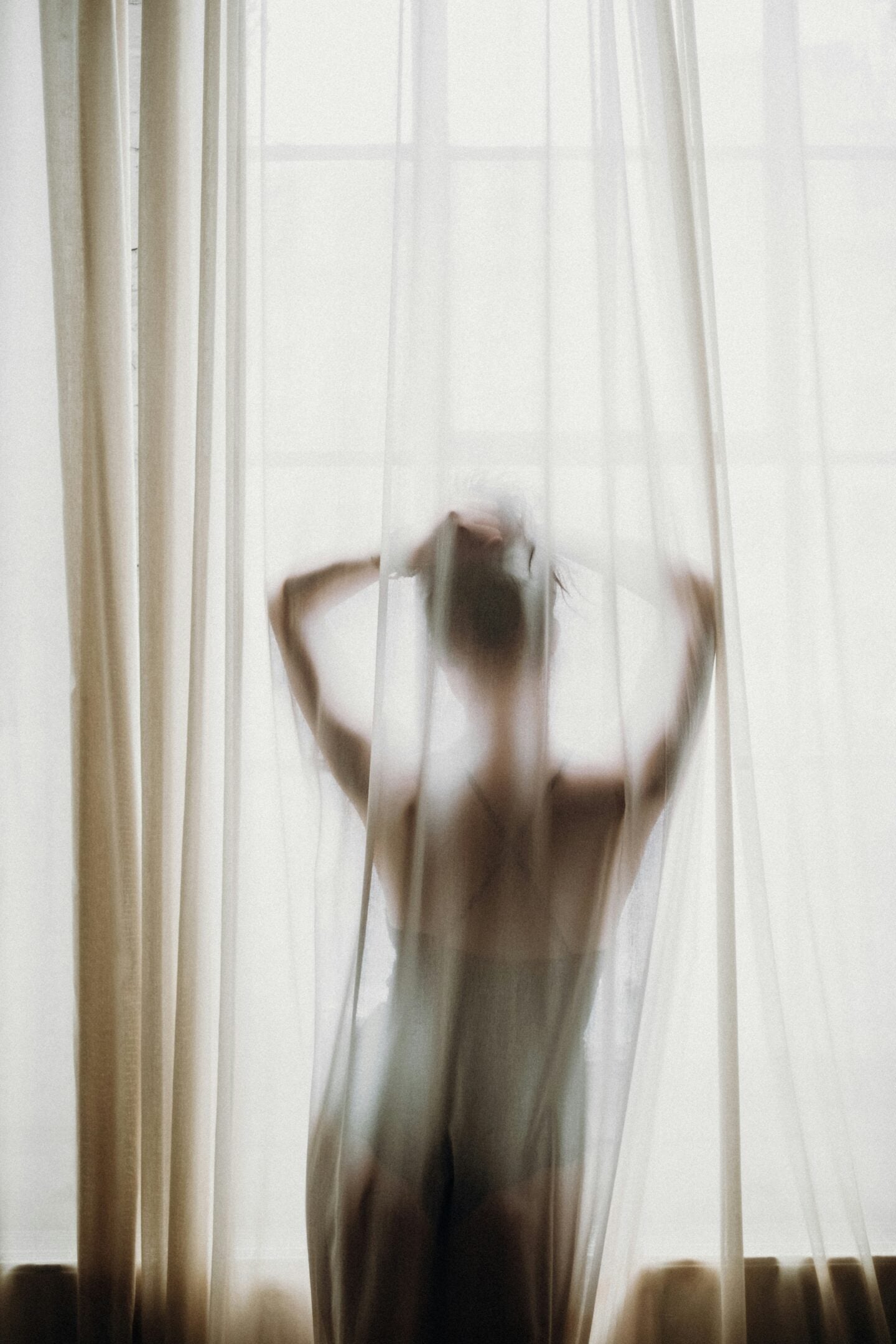 A blurred figure of a woman stands behind white curtains with her arms raised, softly lit by daylight from the window.