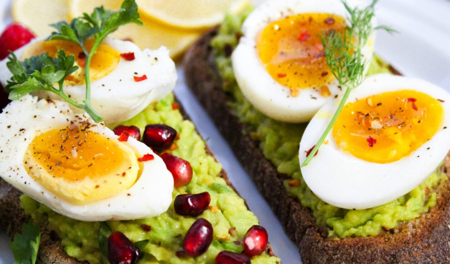 Close-up of avocado toast topped with soft-boiled eggs, pomegranate seeds, fresh herbs, and spices on whole-grain bread.