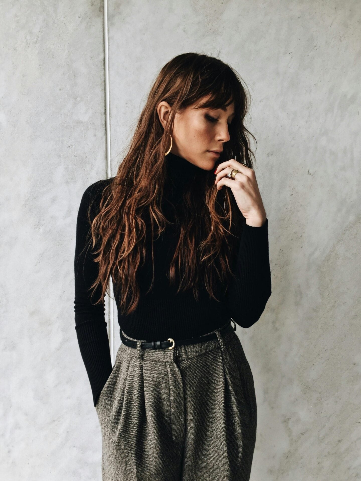 A woman with long wavy hair wears a black turtleneck and high-waisted gray trousers, standing against a textured gray wall with one hand near her face.