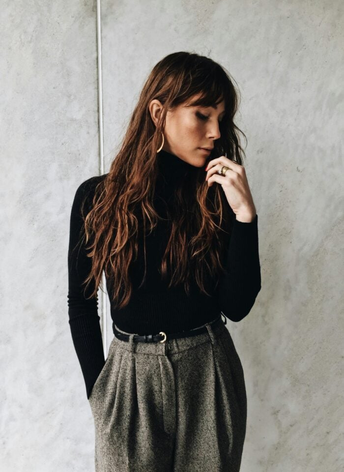 A woman with long wavy hair wears a black turtleneck and high-waisted gray trousers, standing against a textured gray wall with one hand near her face.
