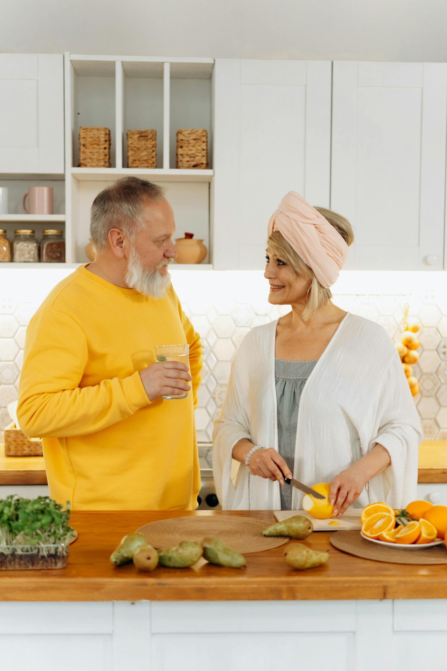 Older couple standing in a bright kitchen, smiling at each other while the woman slices fruit and the man holds a glass of water.