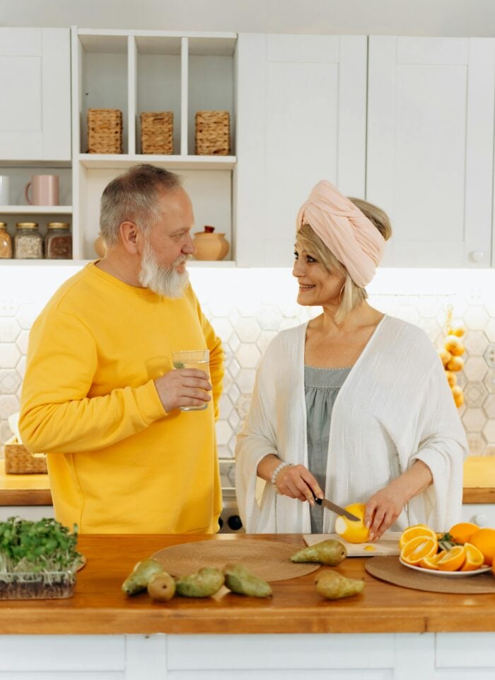 Older couple standing in a bright kitchen, smiling at each other while the woman slices fruit and the man holds a glass of water.