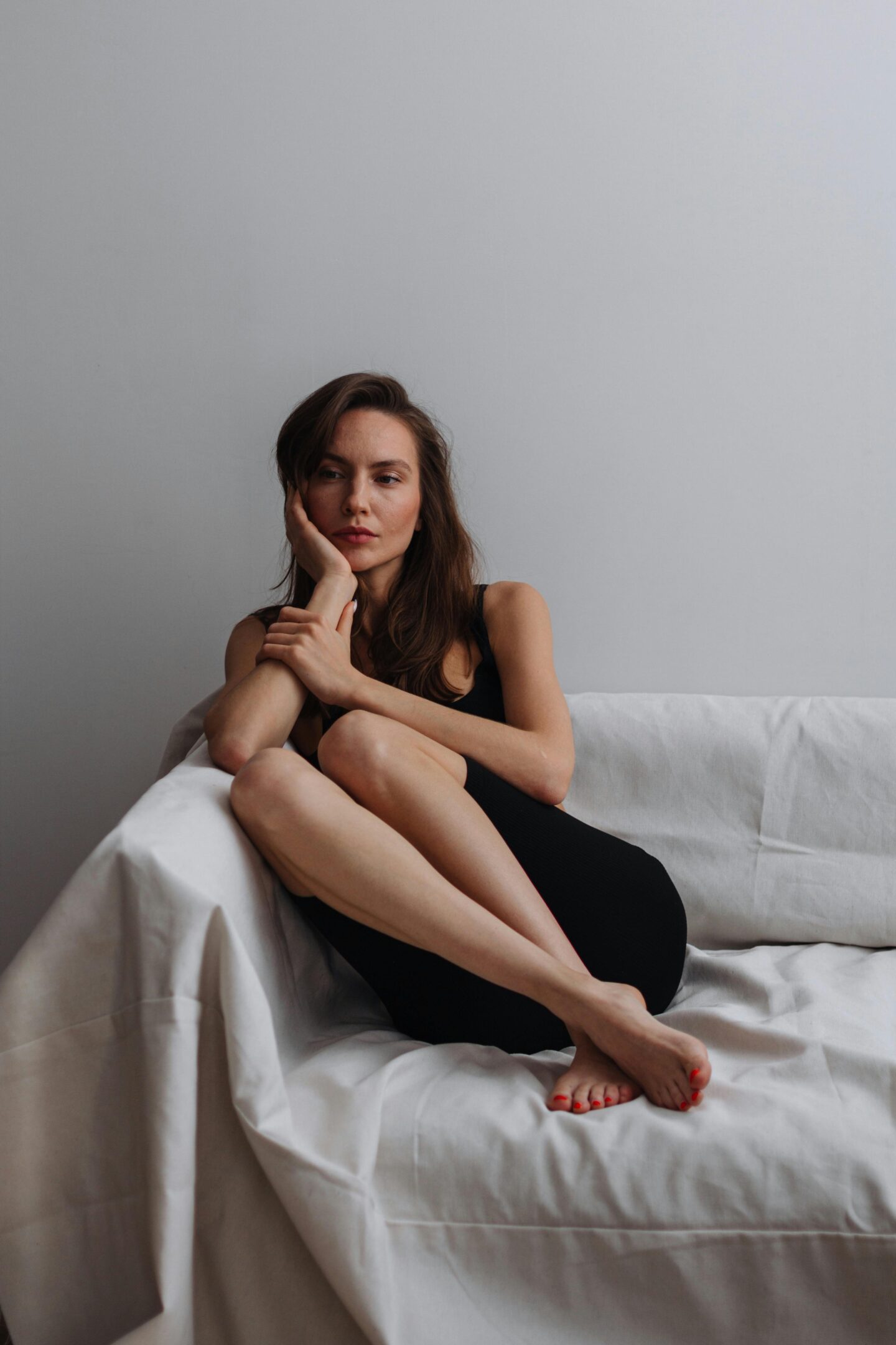 Woman sitting thoughtfully on a white-covered sofa, wearing black shapewear with her legs folded and resting on the seat.