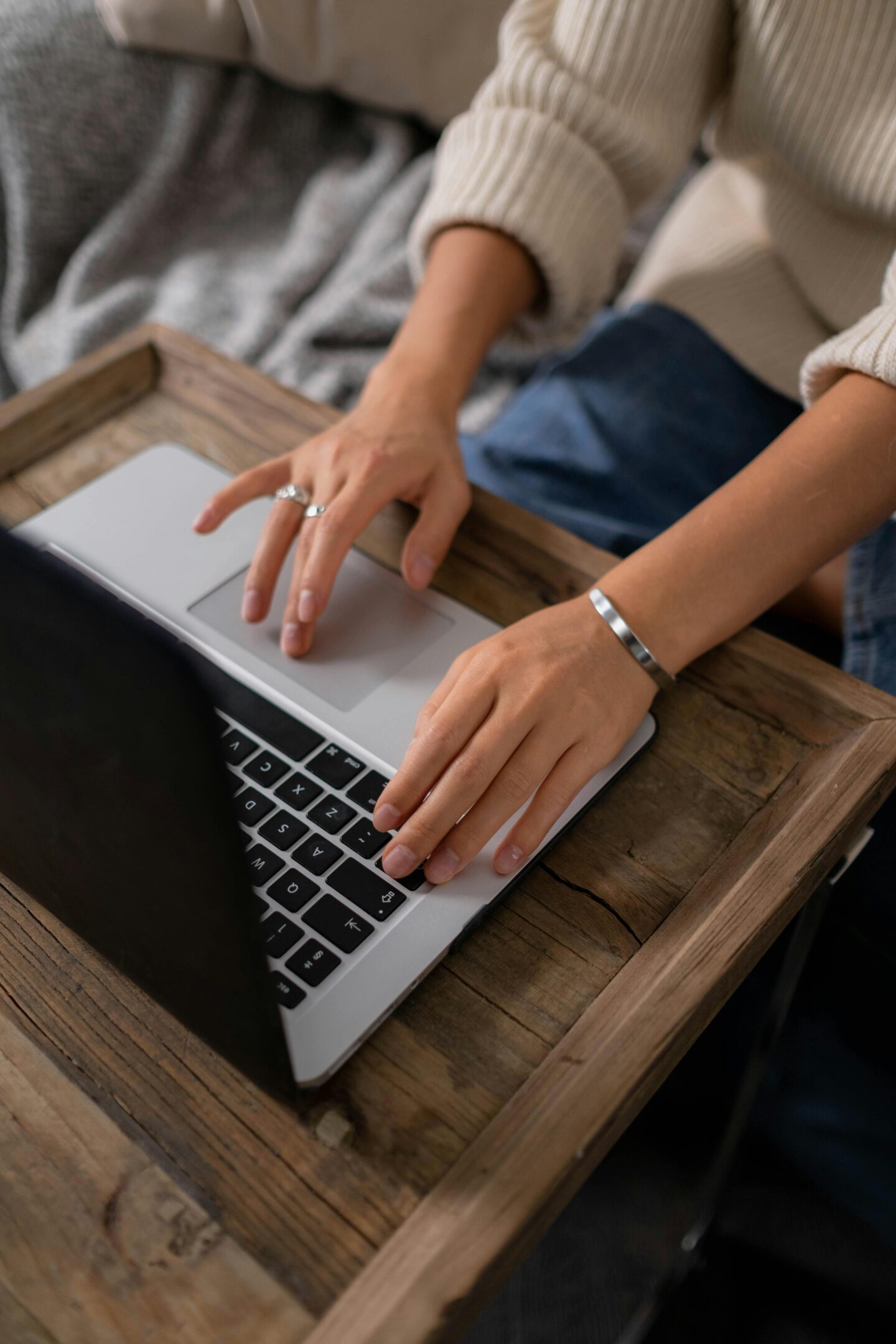 Person typing on a laptop while wearing a silver cuff bracelet and minimalist rings, seated at a wooden table.
