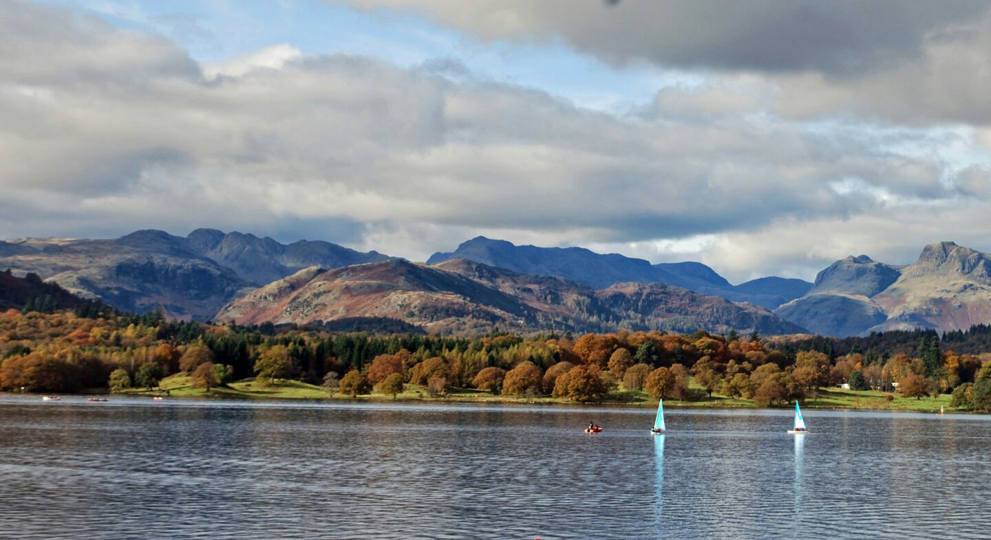 Peaceful view of Lake District mountains and sailboats on the water surrounded by autumn colors.