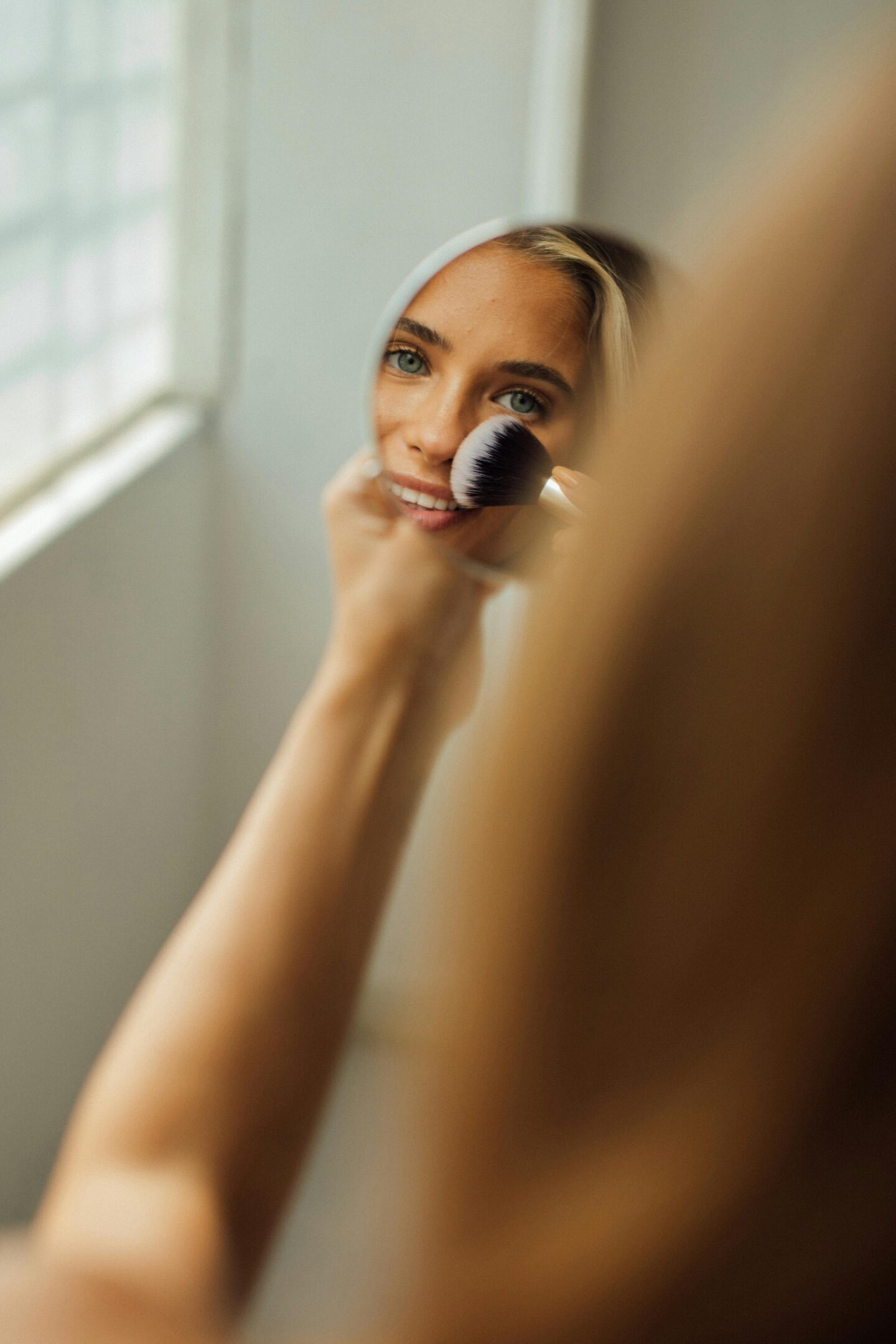 A woman applying makeup with a brush to her nose while looking at her reflection in a small round mirror, illuminated by natural light from a nearby window.
