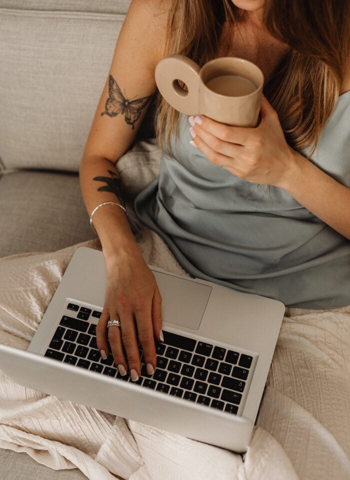 Woman sitting on a sofa with a laptop on her lap, holding a beige coffee mug and wearing a satin top, with visible butterfly and floral tattoos on her arm.