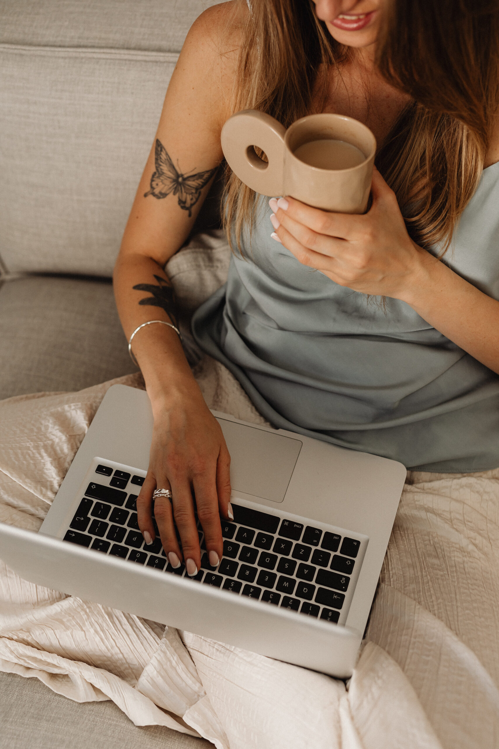 Woman sitting on a sofa with a laptop on her lap, holding a beige coffee mug and wearing a satin top, with visible butterfly and floral tattoos on her arm.