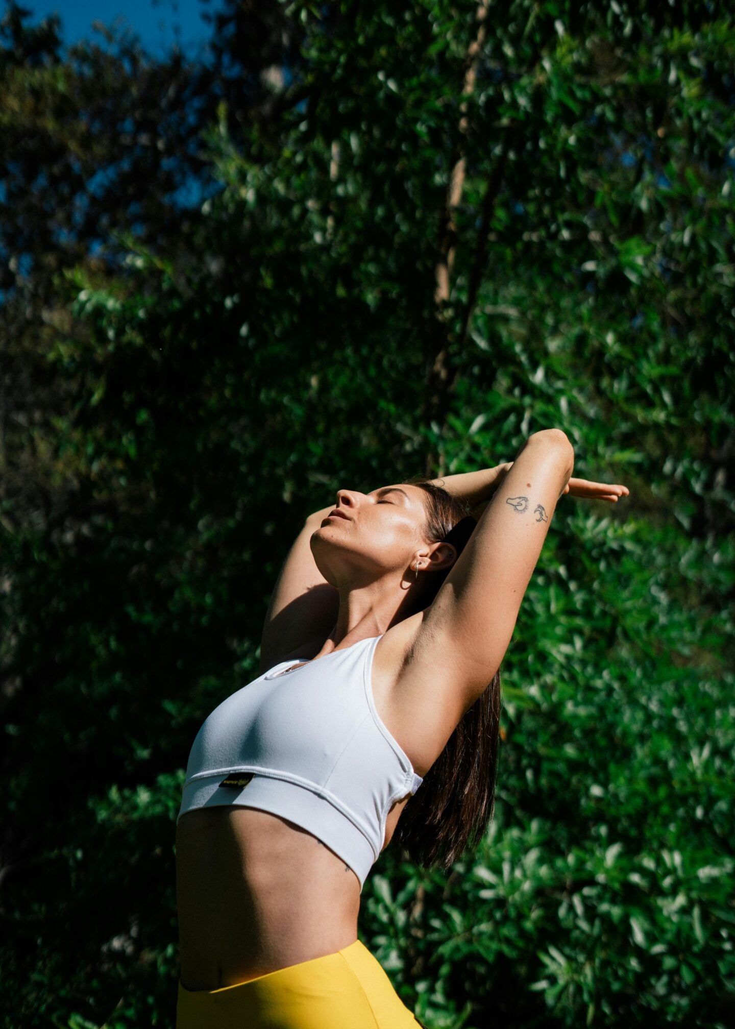 A woman in a white sports bra and yellow leggings stretches with her arms raised above her head, eyes closed, enjoying the sunlight against a background of green trees.