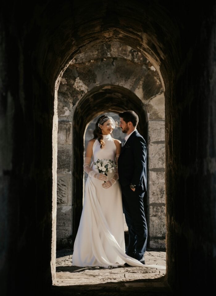 A bride and groom stand closely together inside a dramatic stone archway, the bride holding a bouquet and wearing a long white gown while the groom wears a dark suit.