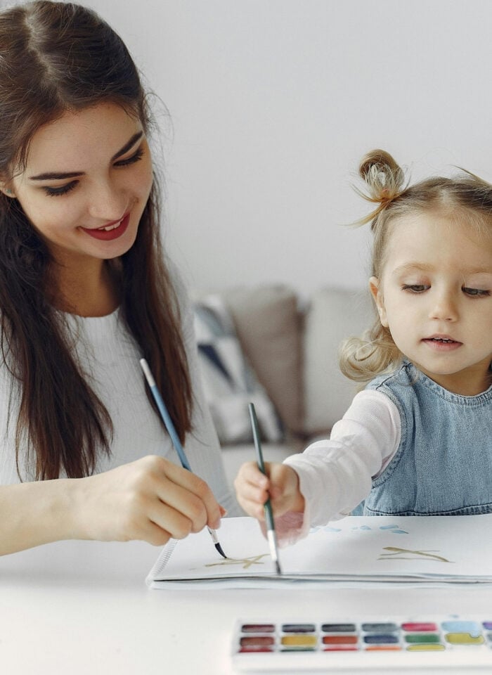 A smiling young woman and a young girl sit at a table painting together with watercolors in a cozy home setting.