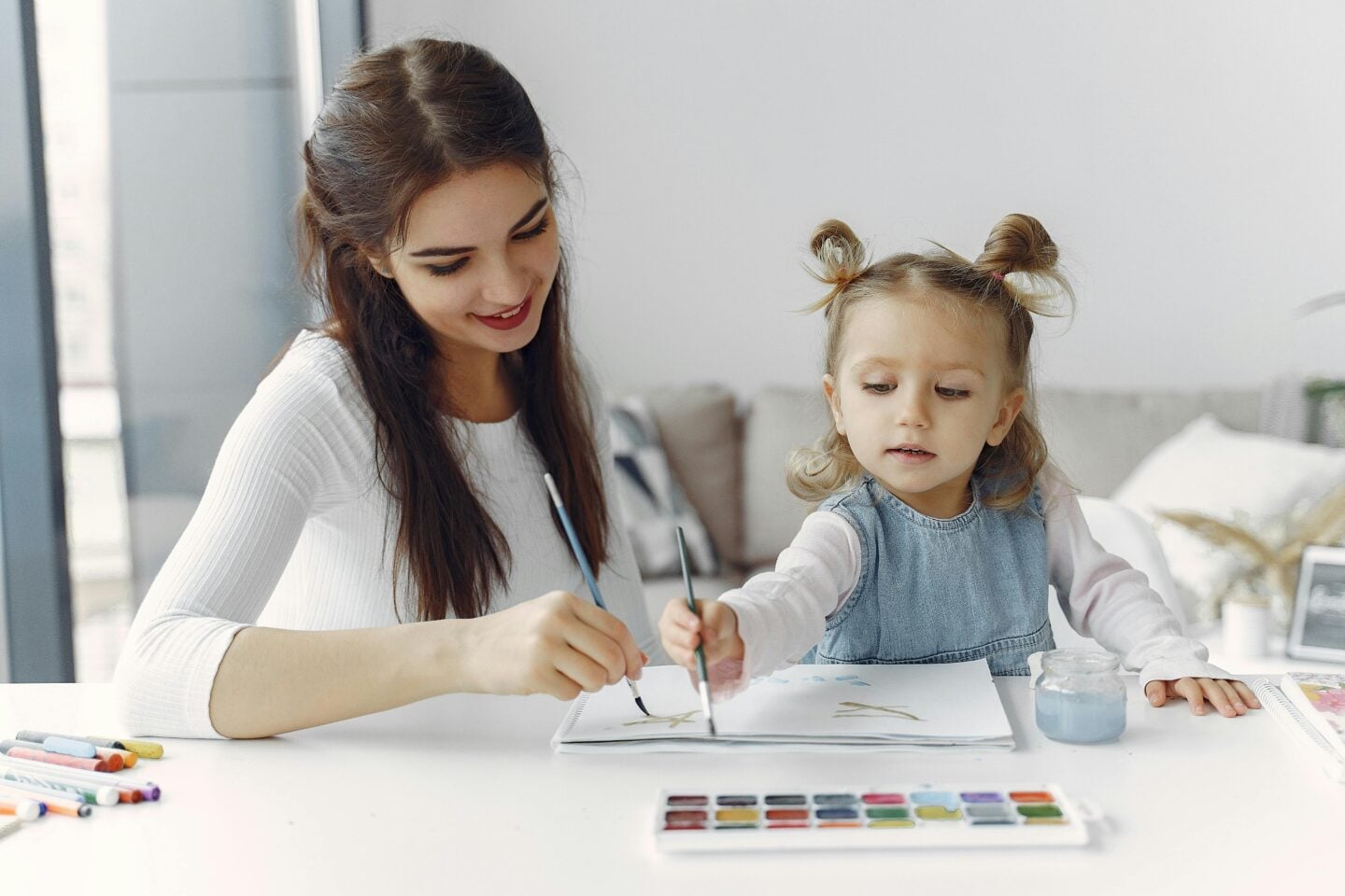 A smiling young woman and a young girl sit at a table painting together with watercolors in a cozy home setting.