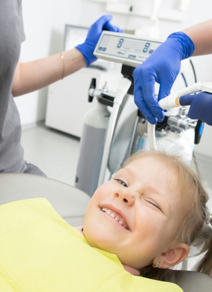 Smiling child receiving dental care with the help of two dentists wearing blue gloves in a pediatric dental clinic.