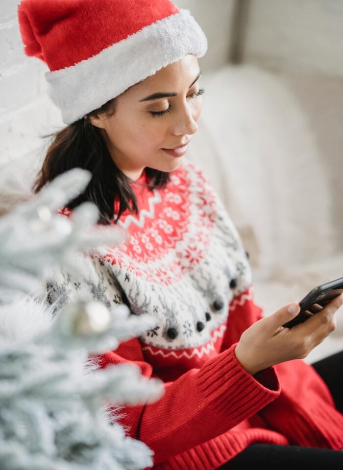 A woman wearing a red Santa hat and a festive holiday sweater sits beside a decorated Christmas tree while looking at her phone.