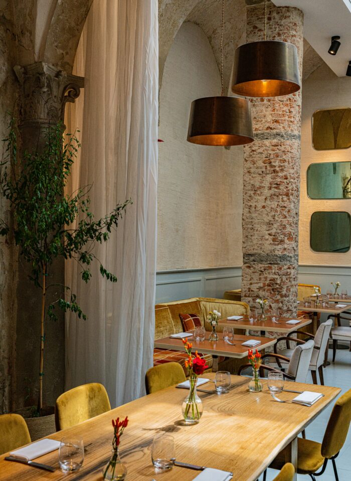 Stylish restaurant interior with long wooden tables, yellow velvet chairs, exposed brick columns, and small flower vases as table decor.