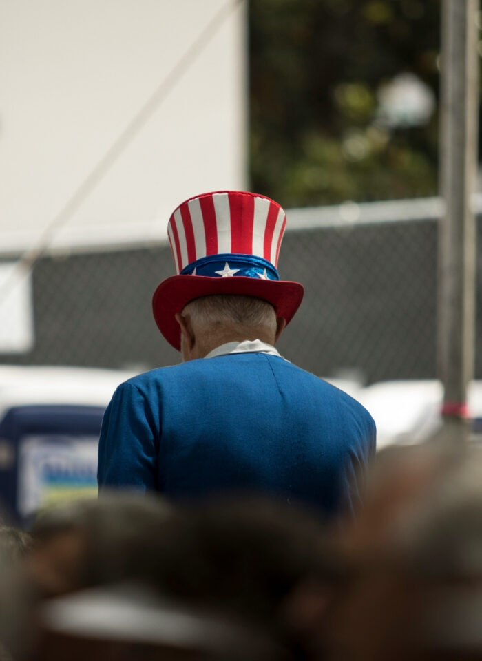 An older man wearing a red, white, and blue Uncle Sam-style hat and blue jacket is seen from behind in a crowded outdoor setting.