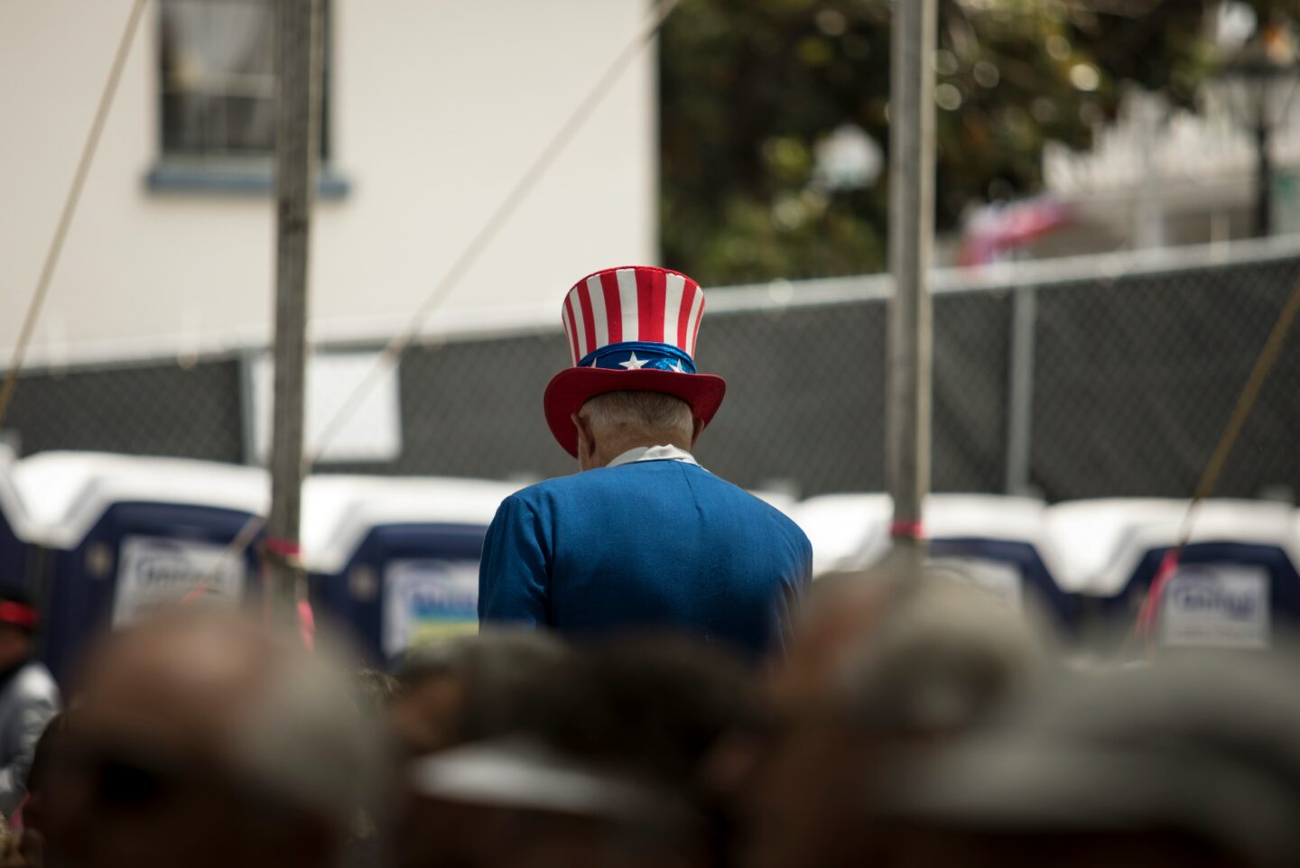 An older man wearing a red, white, and blue Uncle Sam-style hat and blue jacket is seen from behind in a crowded outdoor setting.
