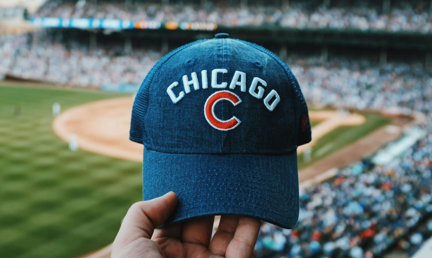 A hand holds up a blue Chicago Cubs baseball cap inside a stadium with the field and crowd blurred in the background.