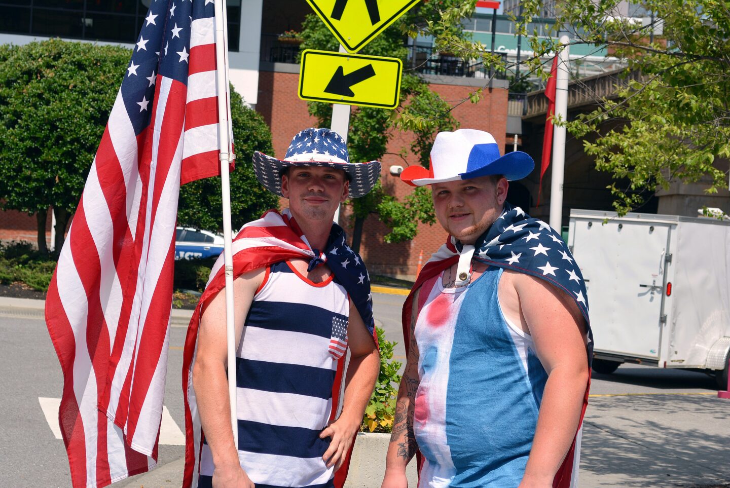Two men dressed in American flag-themed clothing and cowboy hats stand outdoors, one holding a large U.S. flag.