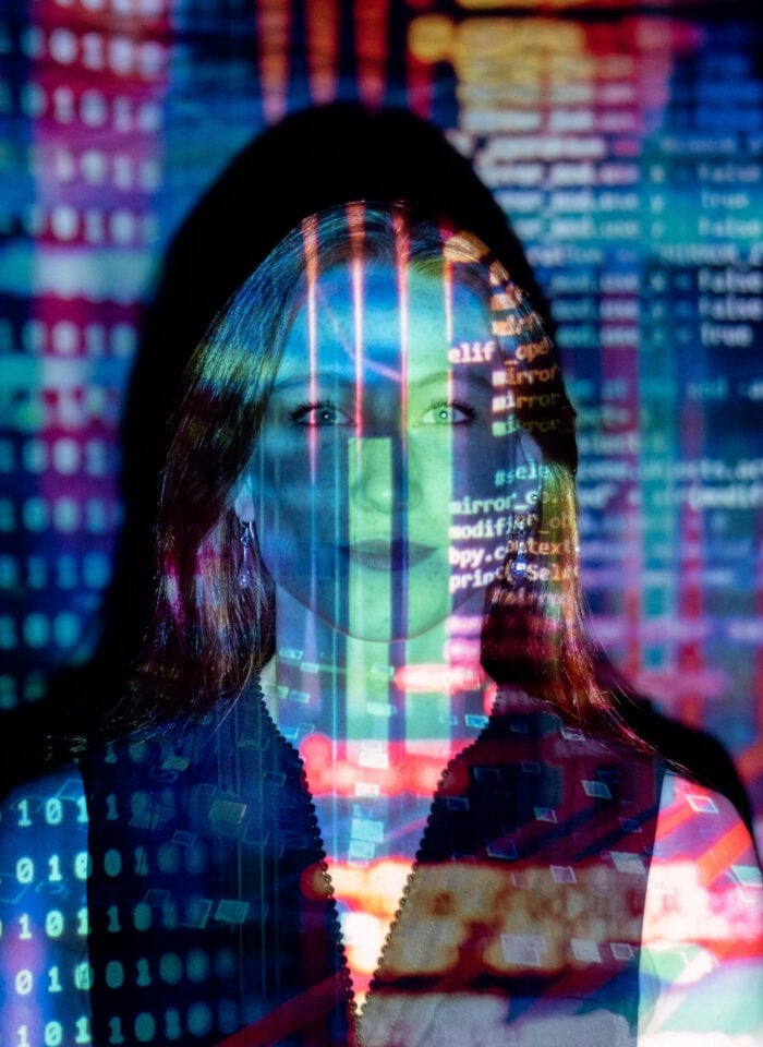 Woman standing in front of a projection of colorful digital code and data patterns, symbolizing technology and cybersecurity.