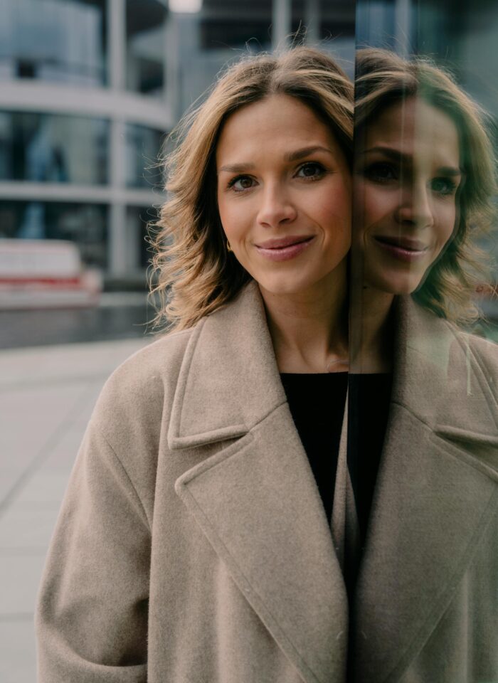 Smiling woman standing outside in a beige coat, reflected in a glass wall.