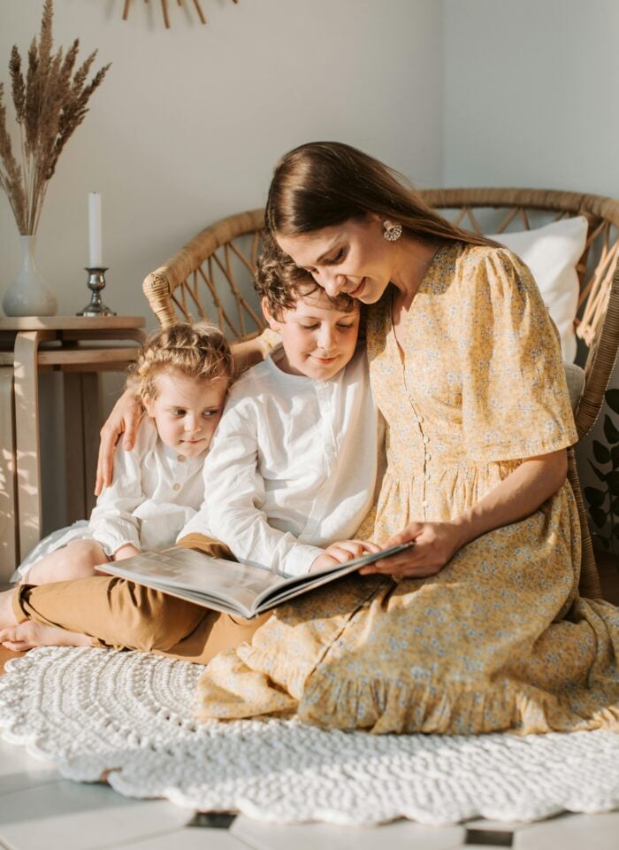 Mother sitting on the floor with her two young children, reading a book together in a bright, cozy living room.