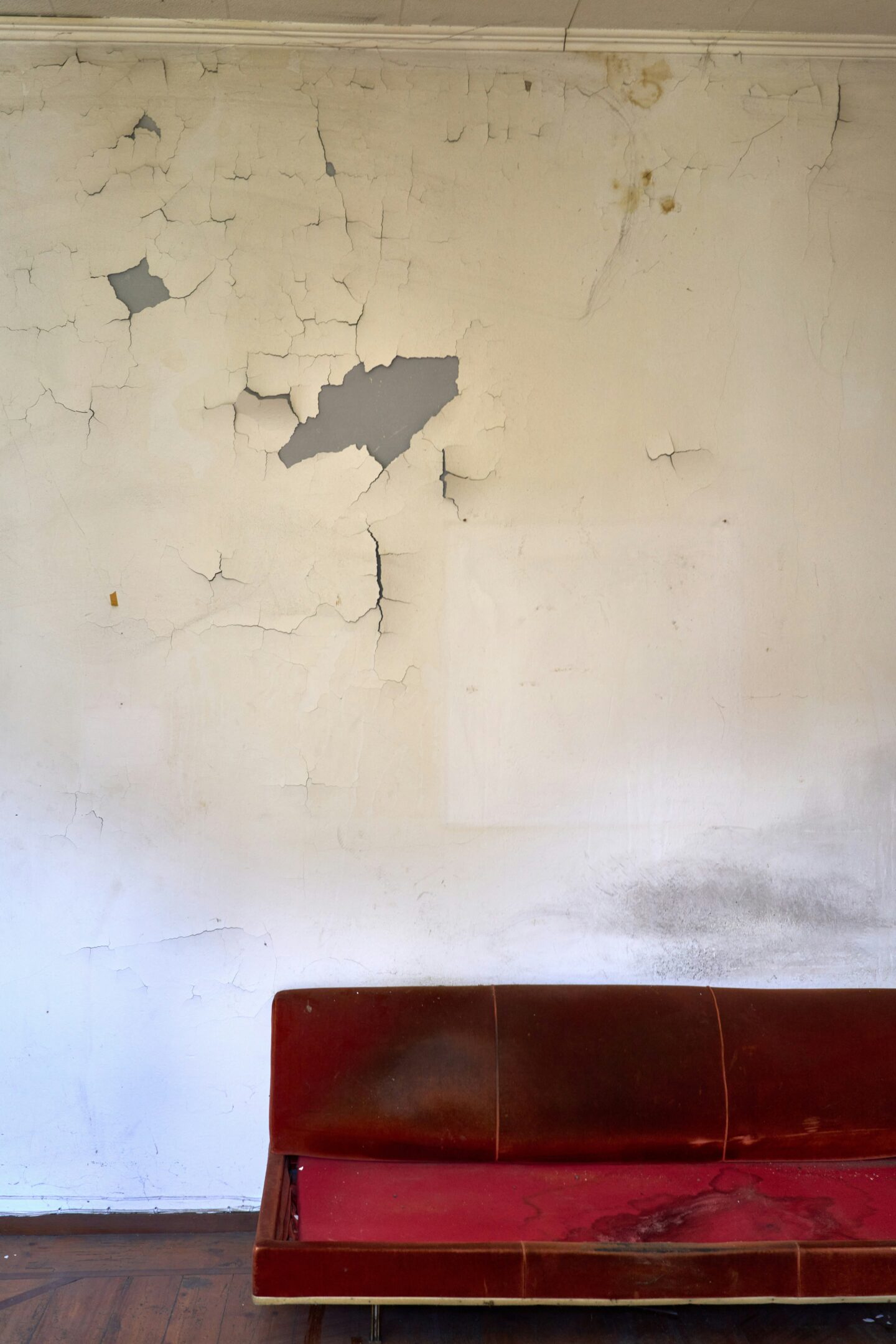 Cracked and peeling interior wall above a worn red sofa, showing visible signs of neglect and damage in a living space.