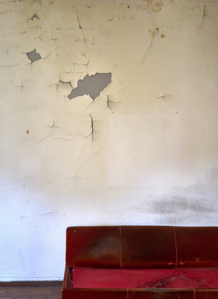 Cracked and peeling interior wall above a worn red sofa, showing visible signs of neglect and damage in a living space.