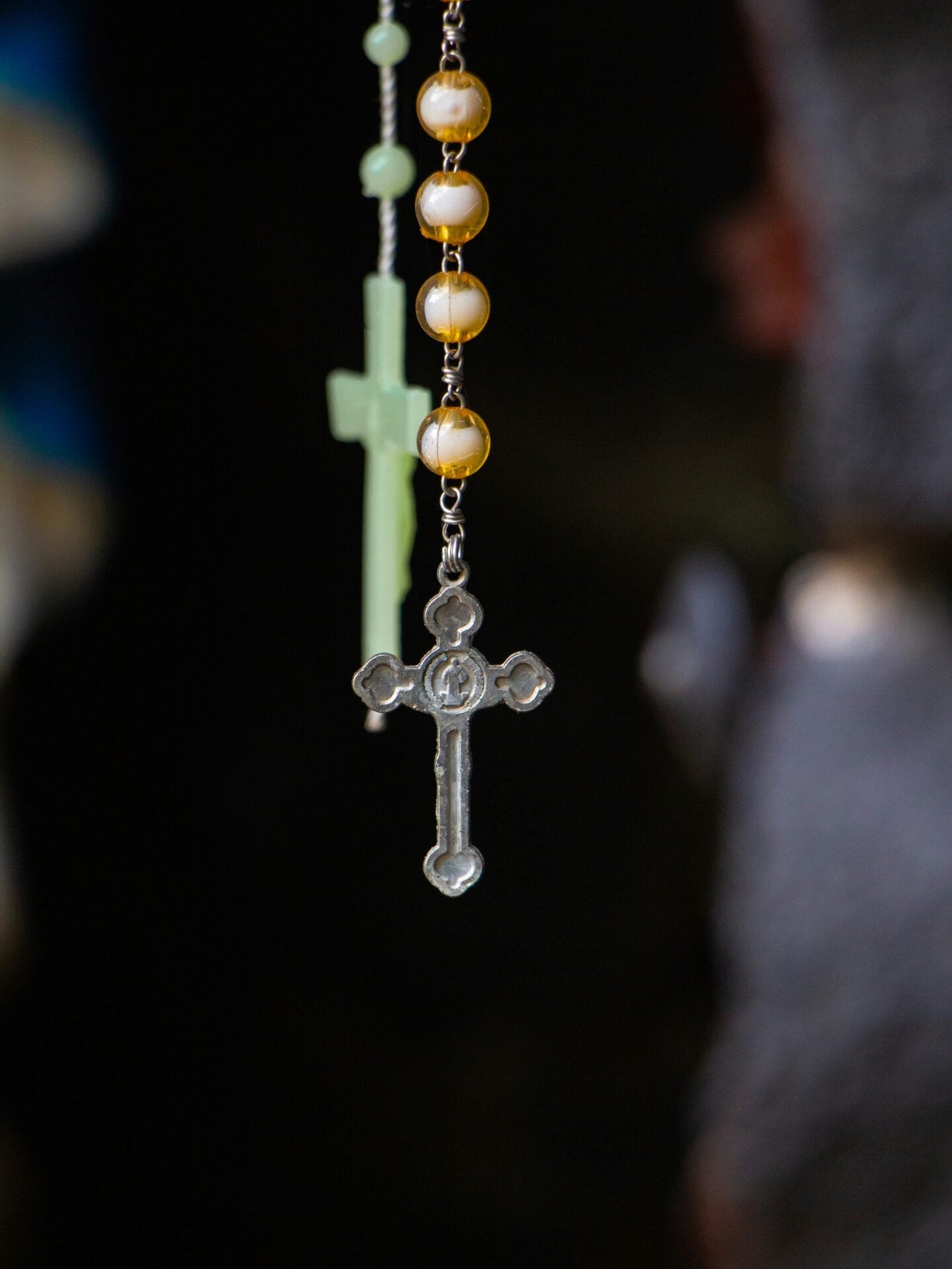 Close-up of a rosary with pearl-like beads and a small metal crucifix hanging in focus against a dark background.