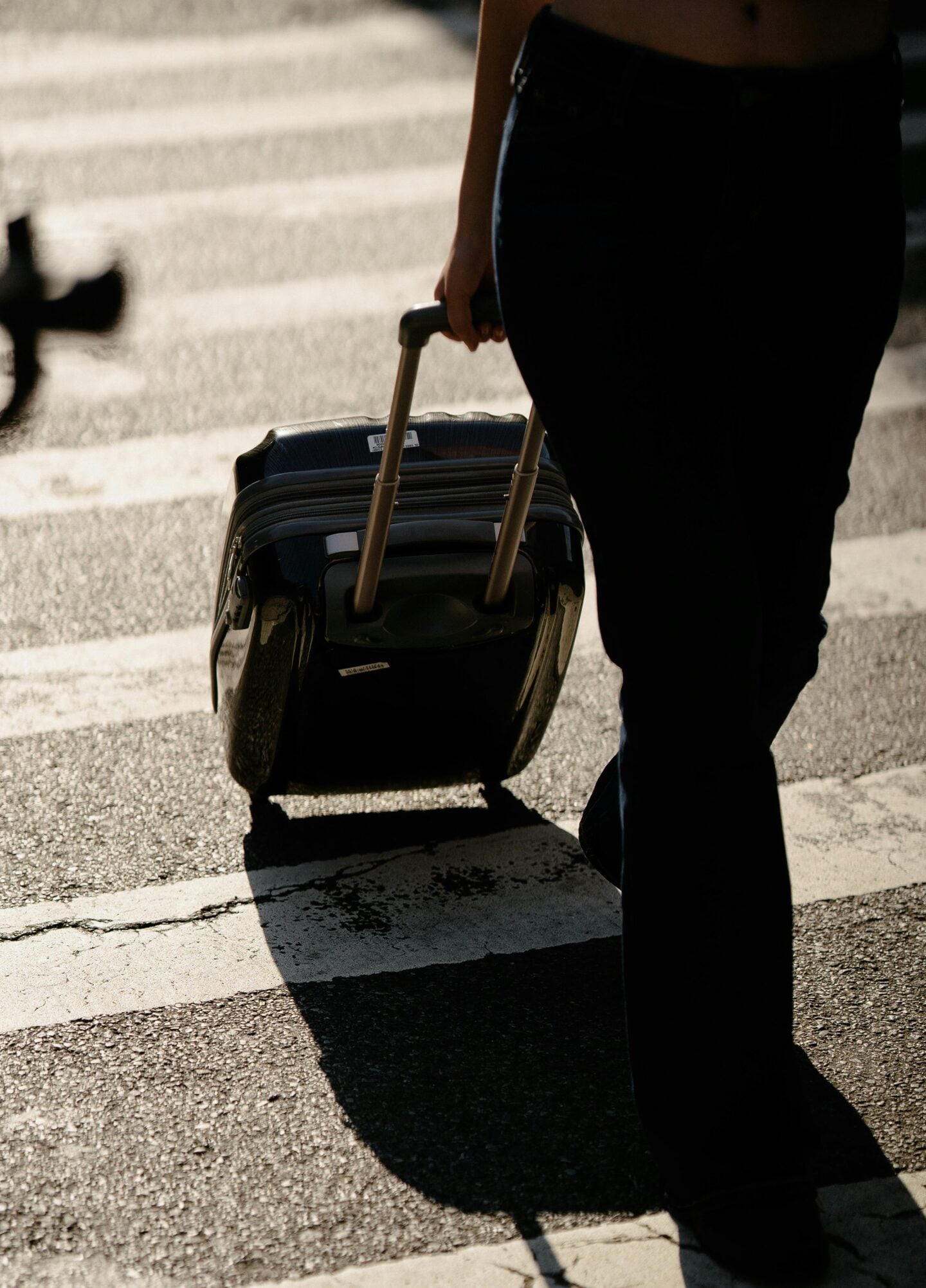 Traveler walking across a street with a small wheeled suitcase, sunlight casting long shadows on the pavement.