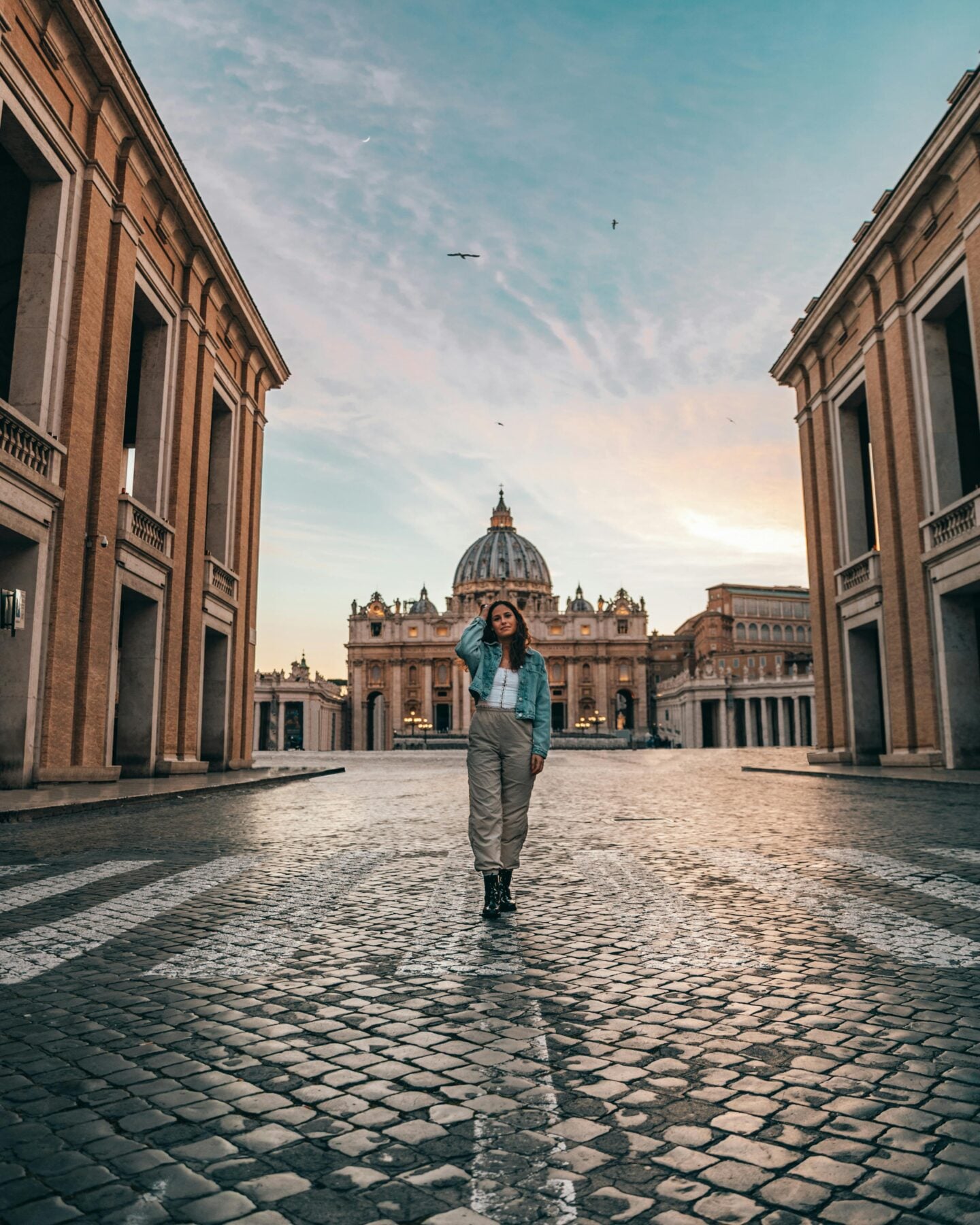 Woman walking down an empty cobblestone street in front of St. Peter’s Basilica in Vatican City during sunrise.