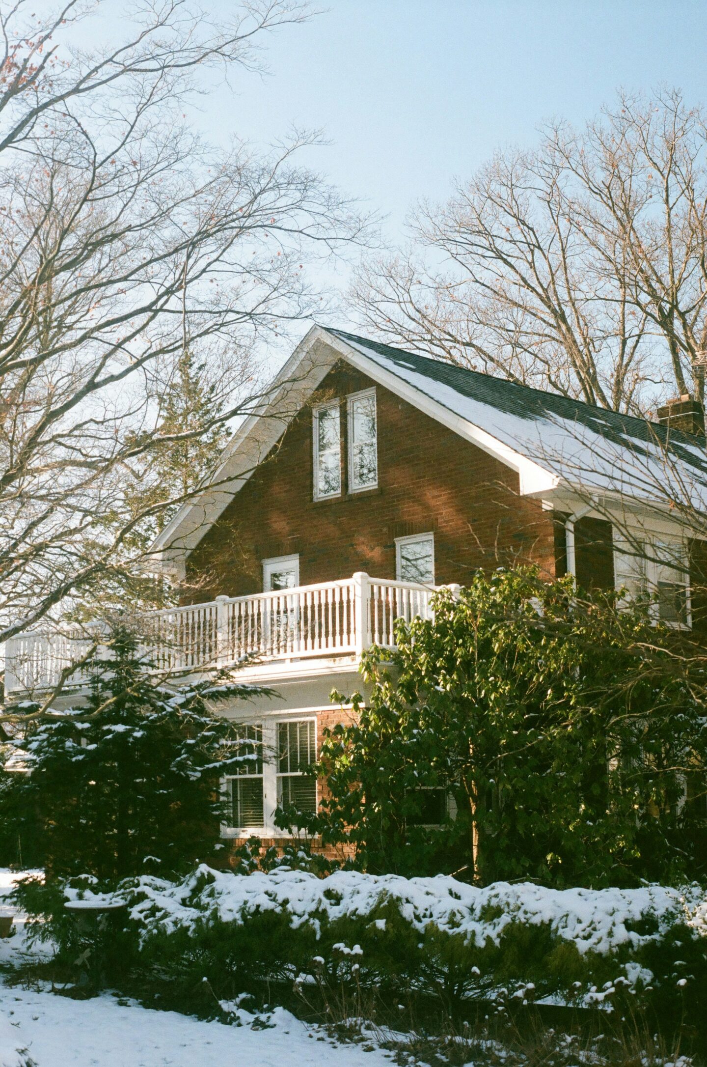 Two-story brick house surrounded by snow-covered trees and shrubs on a clear winter day.