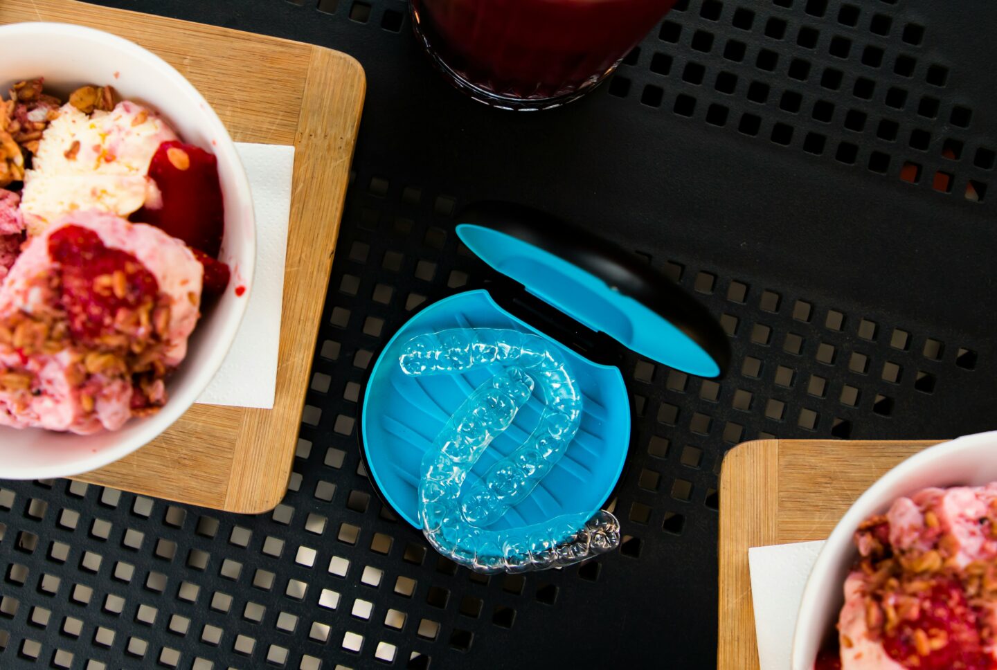 A blue case with clear dental aligners sits on a black table beside bowls of yogurt with fruit and granola and a glass of juice.