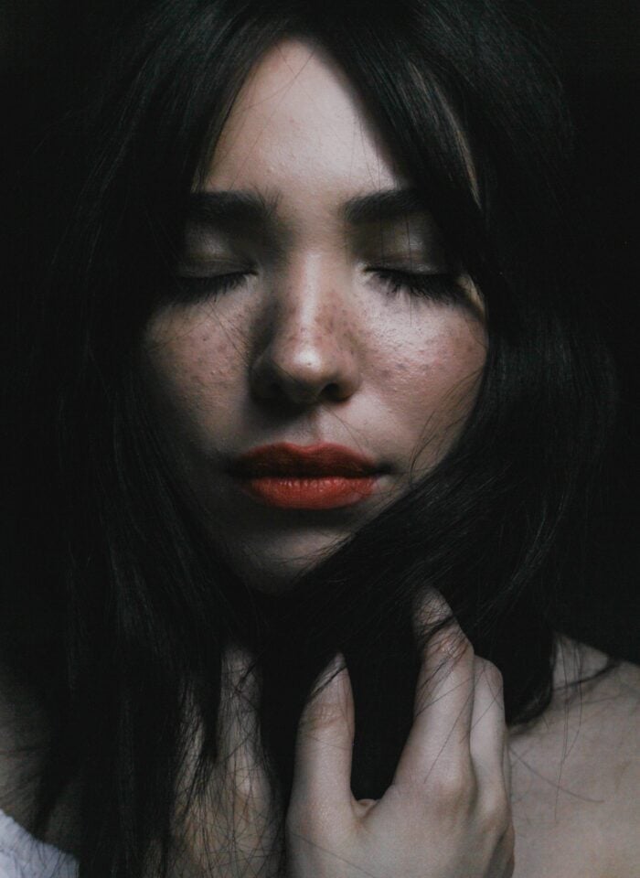 Close-up portrait of a woman with closed eyes and red lipstick, gently touching her face in dim lighting.
