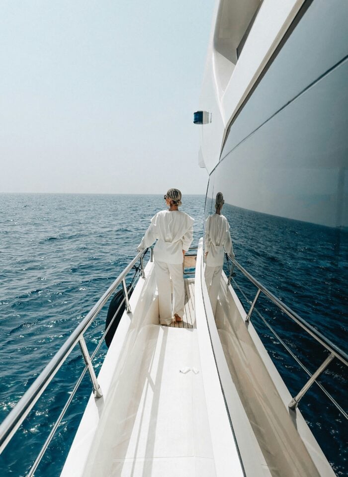 A person dressed in a light outfit stands barefoot on the side deck of a luxury yacht, looking out at the calm open sea on a bright, clear day.