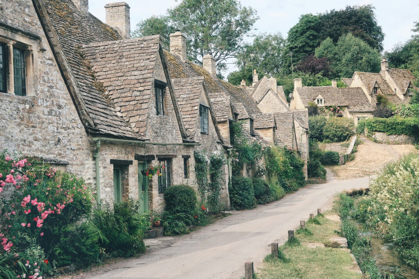A row of traditional stone cottages in the Cotswolds, surrounded by greenery and flowers, with a narrow country lane running alongside them.
