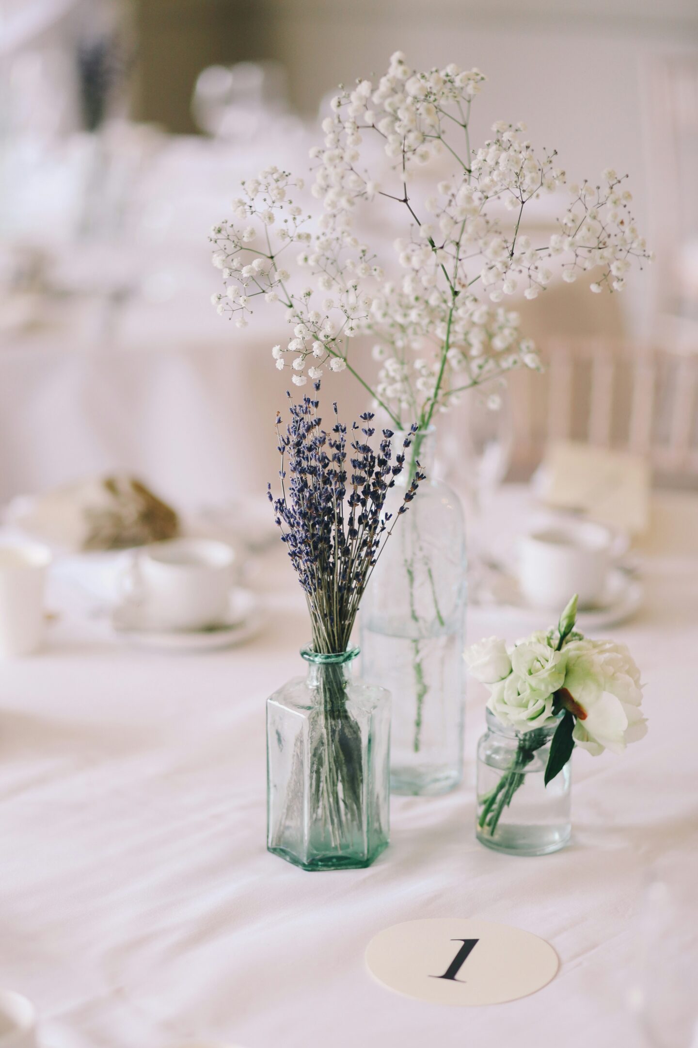 A soft, elegant table centerpiece featuring dried lavender, baby’s breath, and white flowers arranged in small glass bottles on a white tablecloth.
