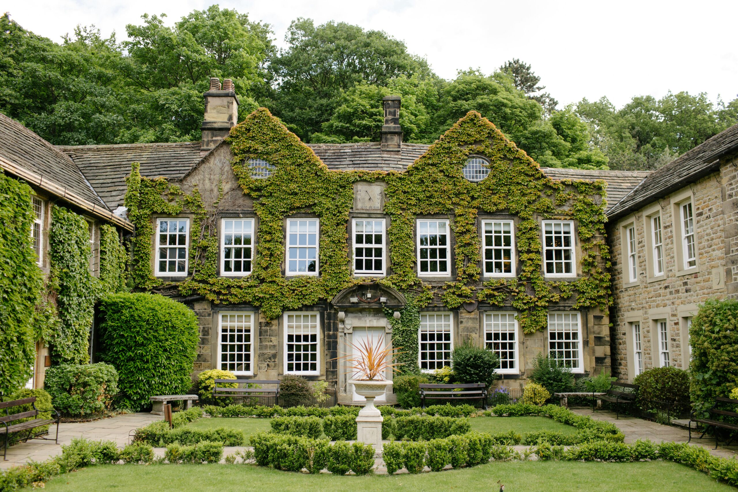 An elegant historic building covered in green ivy, surrounded by manicured gardens and trees, creating a picturesque outdoor wedding venue setting.