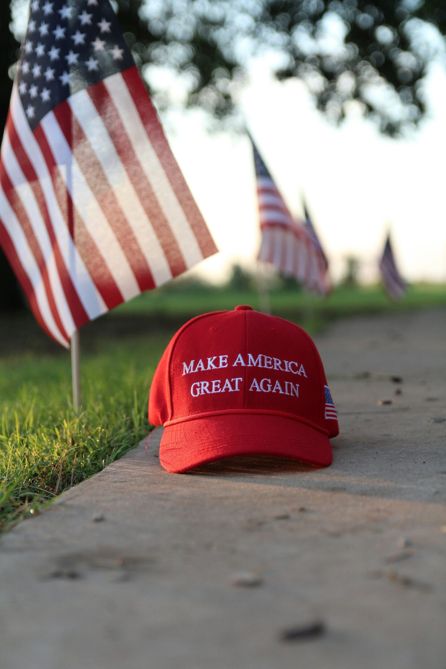A red “Make America Great Again” hat sits on a sidewalk surrounded by small American flags planted in the grass.