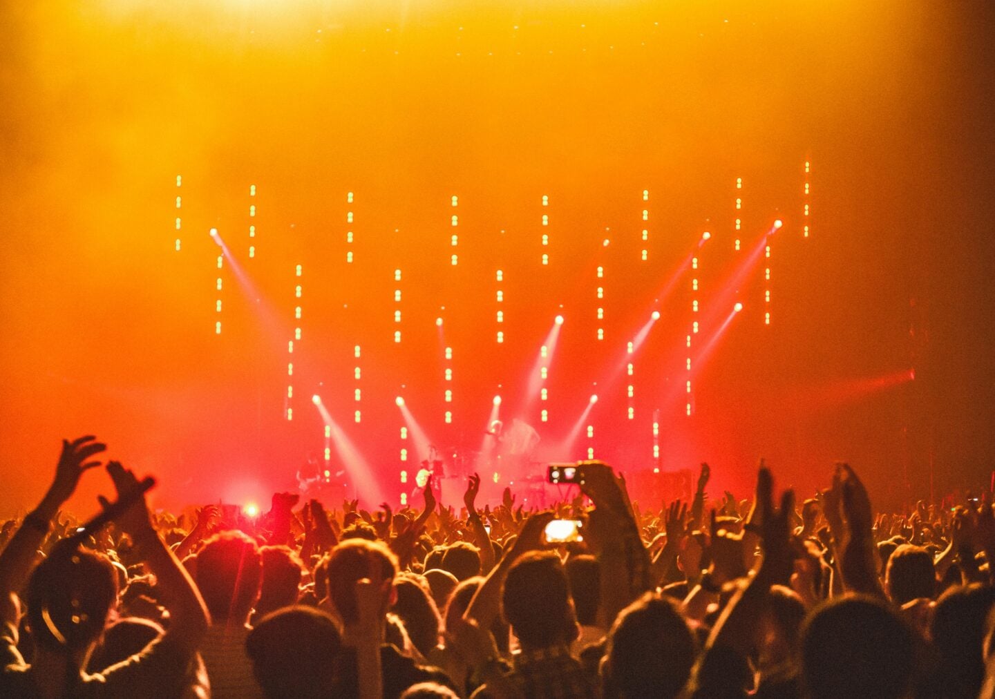A large crowd of people with raised hands enjoying a live concert, with bright orange and yellow stage lights shining down on the performers in the background.